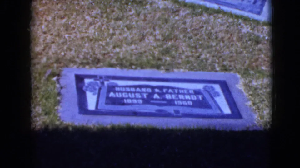 Final 8mm frame holds on the August A. Bernot grave marker in the grassy cemetery, with visible grain and cool color shift.