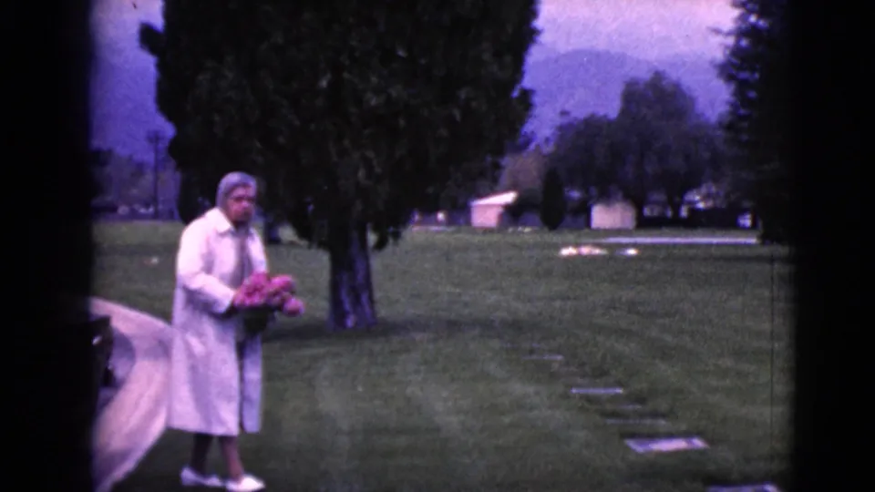 Older woman in a light coat carries pink flowers through a 1968 Clarksdale, Arizona cemetery scene.
