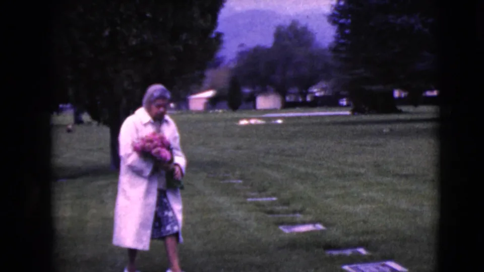 Older woman holding pink flowers moves among flat cemetery markers in the supported 1968 Clarksdale, Arizona footage.