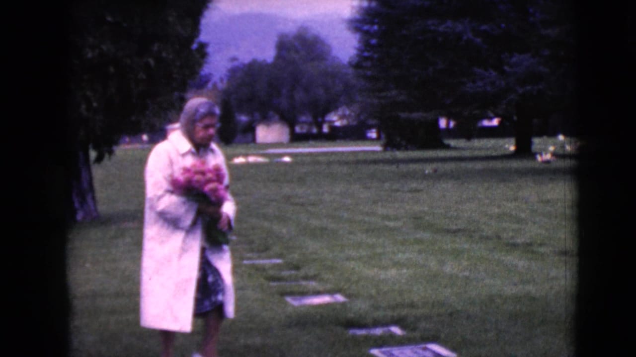 In 1968 Clarksdale, Arizona footage, a woman in glasses walks through a cemetery holding a bouquet of pink flowers.