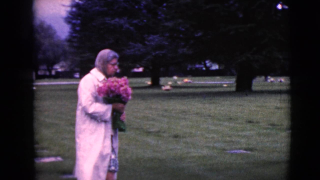 Woman in a white coat holds pink flowers while moving across cemetery grass.