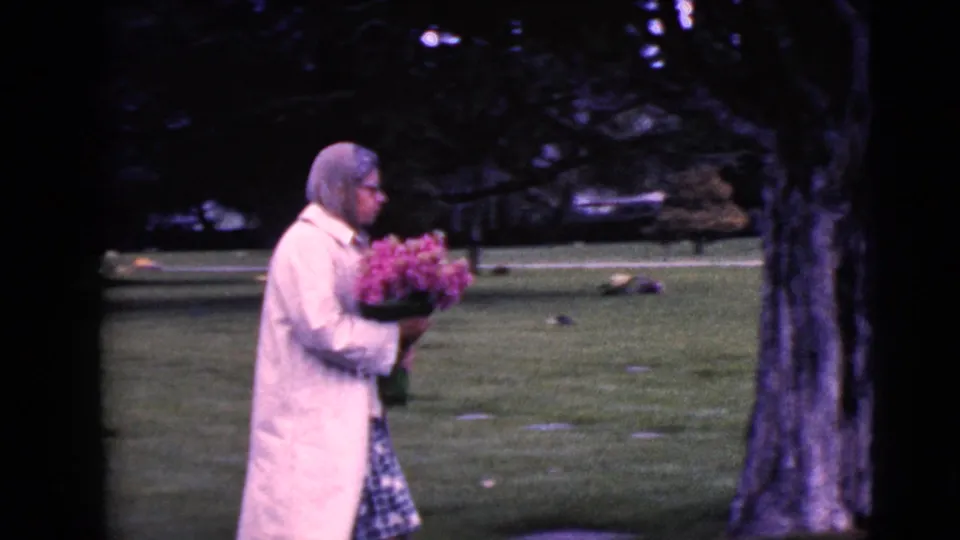 A woman in a light coat and glasses carries flowers past a tree and low grave markers in the cemetery.