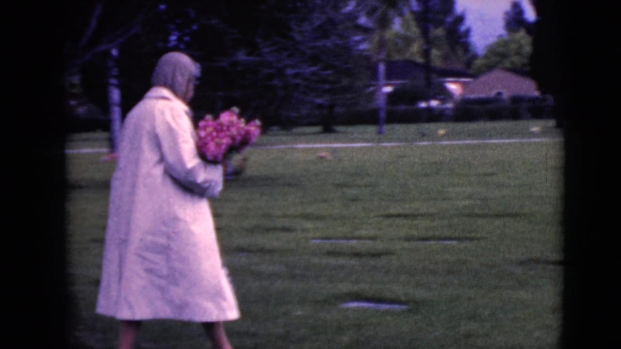Woman in a pale coat carries flowers across a grassy cemetery.