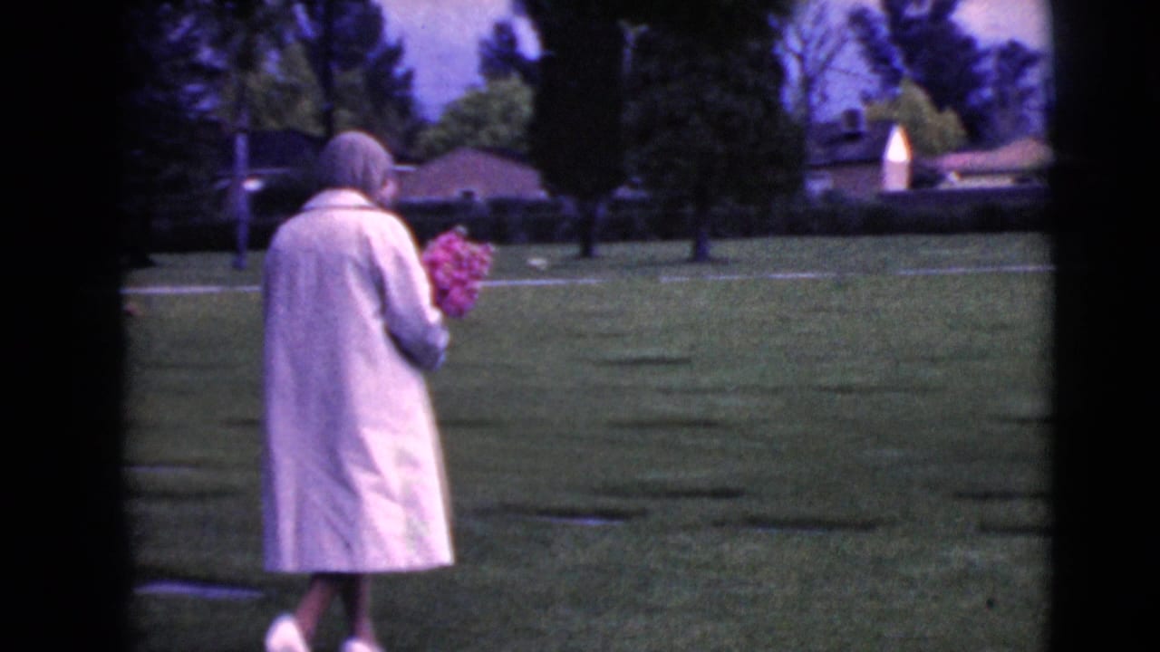 The woman continues through the Clarksdale cemetery, seen from behind with a bouquet and rows of low grave markers in the grass.