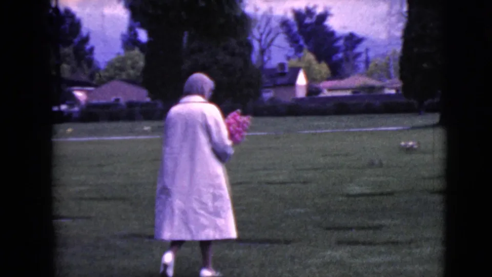 A woman in a pale coat walks farther into the grassy Clarksdale cemetery, holding flowers as grave markers lie scattered ahead.