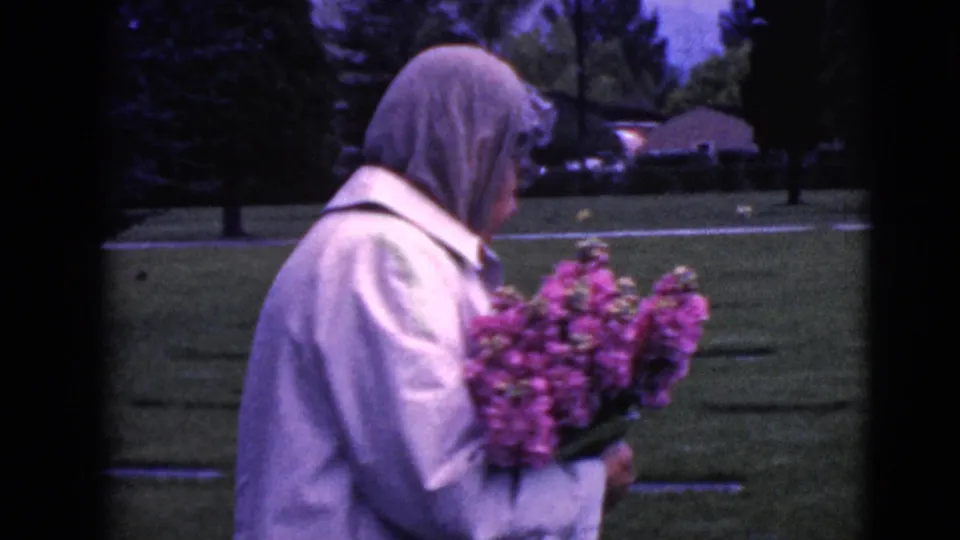 A closer 1968 home-movie frame shows the woman in profile holding a large pink-purple bouquet beside cemetery markers.
