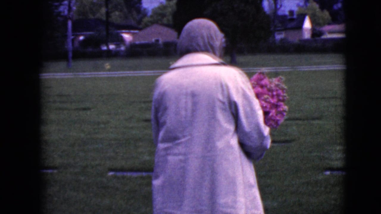 The woman continues through the cemetery grounds with a bouquet, with trees, grass, and houses beyond the graves.