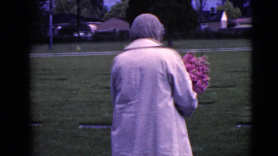 The woman continues through the cemetery grounds with a bouquet, with trees, grass, and houses beyond the graves.