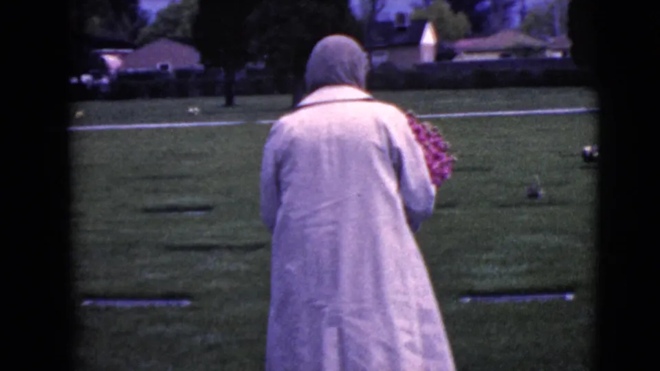 A rear view shows the woman moving past low grave markers in the grassy cemetery, bouquet held close to her body.