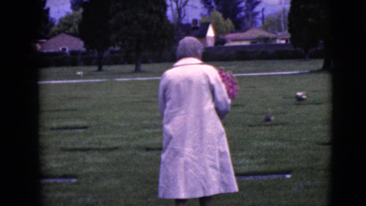 Older woman in a light coat walks across a cemetery lawn holding flowers.
