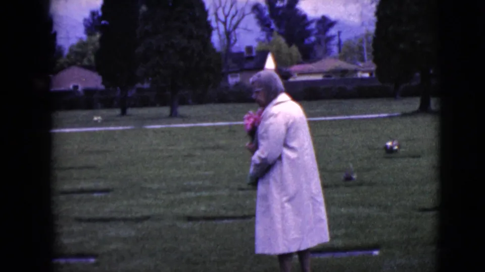 Side view of the older woman in a light coat holding flowers in a Clarksdale cemetery setting.