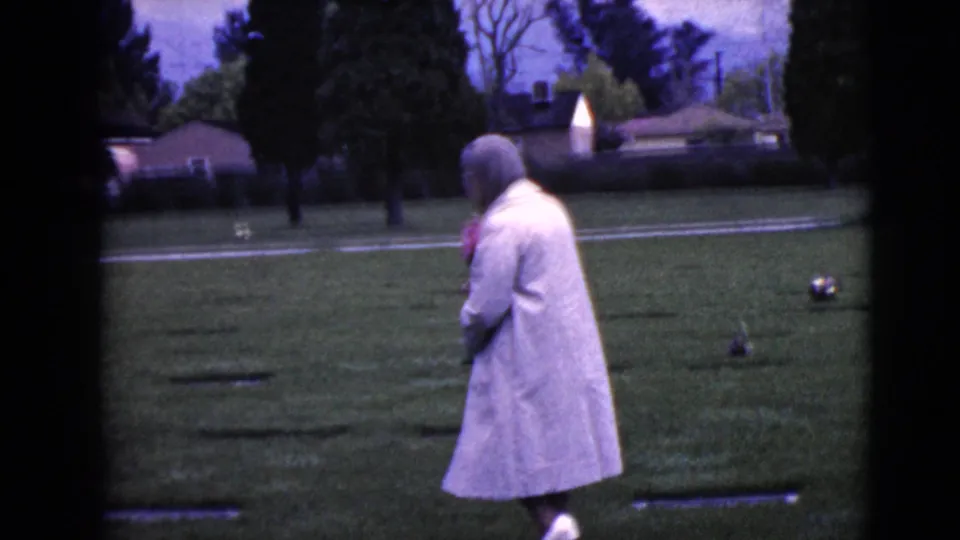 The woman, partly in profile, walks across the cemetery lawn with flowers near low grave markers.