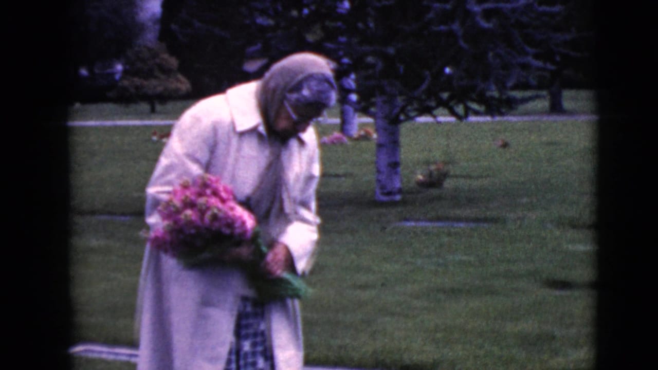 Elderly woman holds a bouquet while standing near a grave marker in a cemetery.