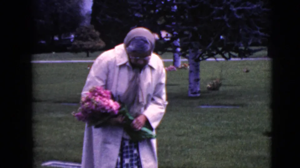 The woman leans forward with a bouquet in hand, appearing to examine or arrange flowers near a grave marker.
