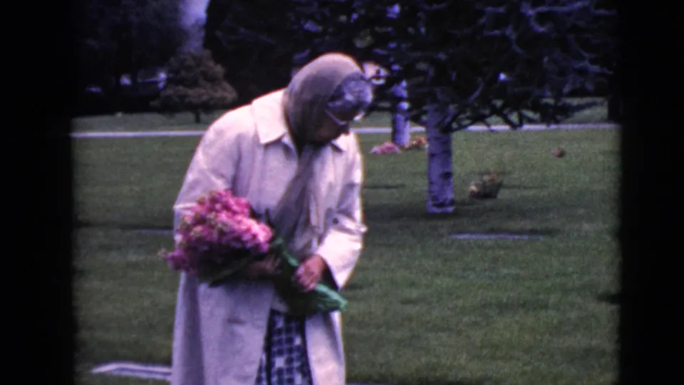At the end of the clip, the woman remains bowed near the grave marker, holding a bouquet on the cemetery lawn.