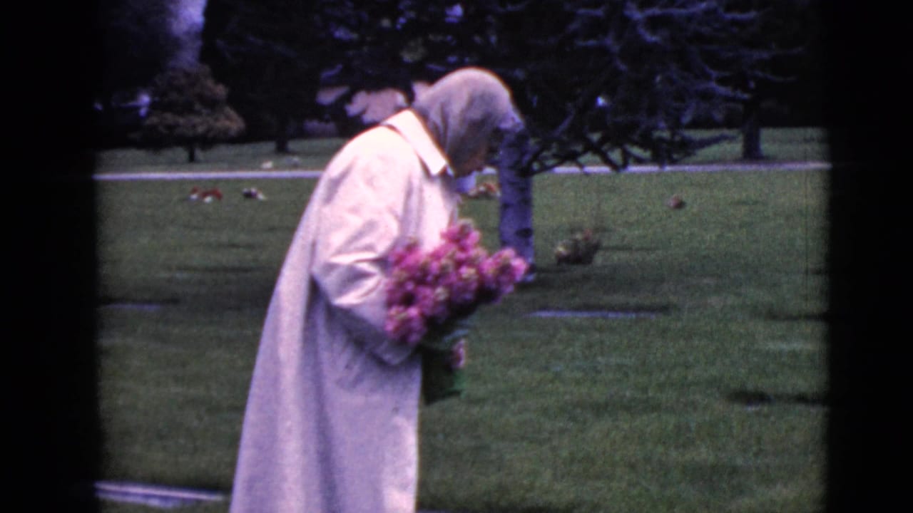 Older woman in a pale coat carries pink flowers through a grassy cemetery.