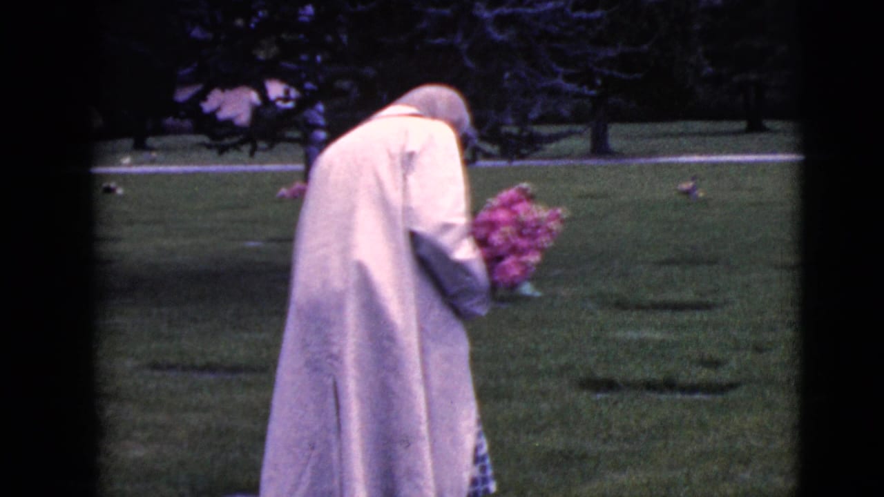 The woman continues through the Clarksdale cemetery with her head bowed, holding a bouquet near low grave markers.
