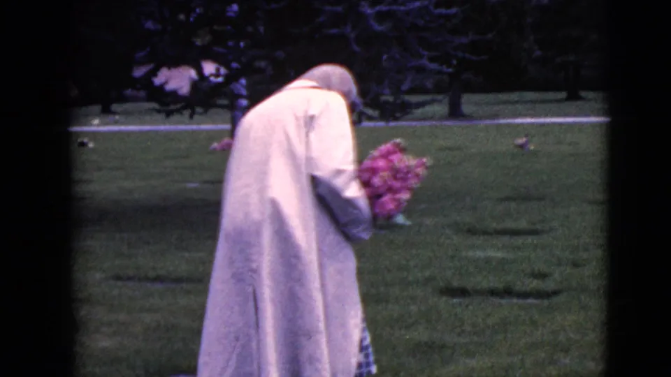 The woman continues through the Clarksdale cemetery with her head bowed, holding a bouquet near low grave markers.