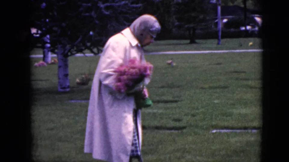 A closer 1968 home-movie frame shows the woman holding pink flowers while standing among cemetery markers and trees.