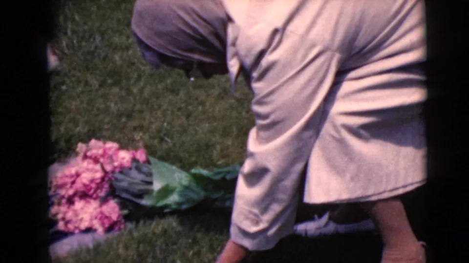 The woman bends toward the cemetery grass to set a bouquet of pink flowers beside a grave marker in the 1968 Arizona home movie.