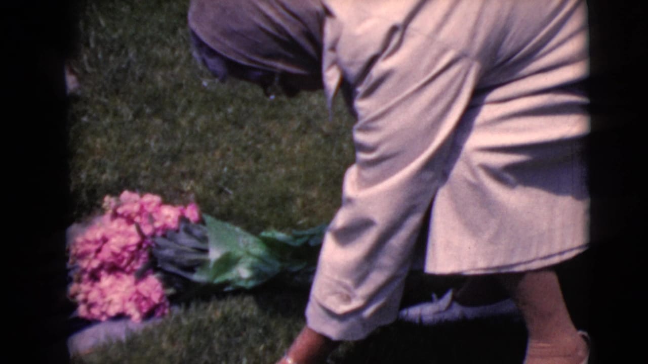 Woman in a pale coat bends near pink flowers and a grave marker on cemetery grass.