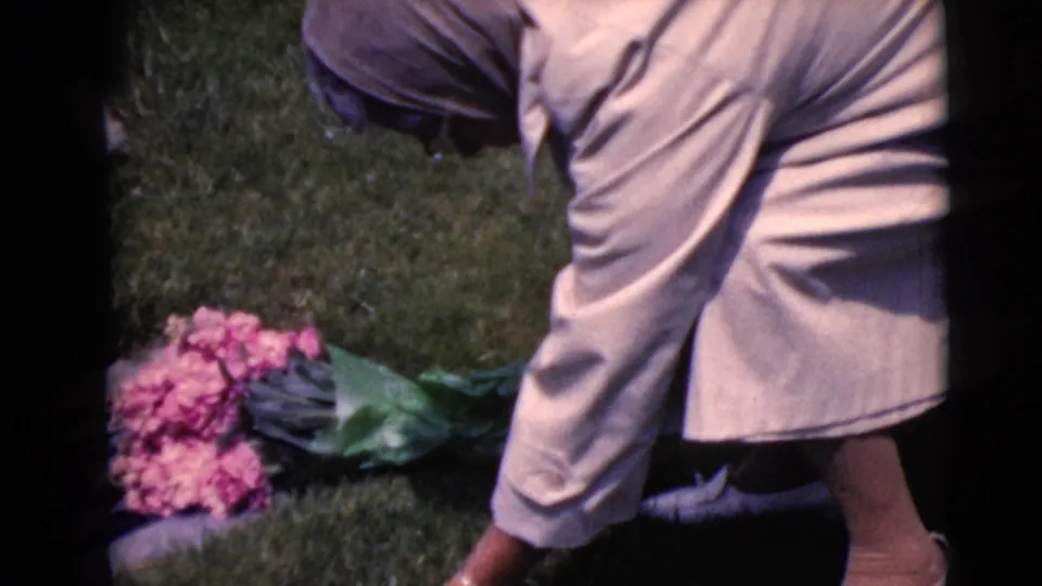 In 1968 Clarksdale, Arizona home-movie footage, a woman bends toward a grave area with pink flowers on the grass.