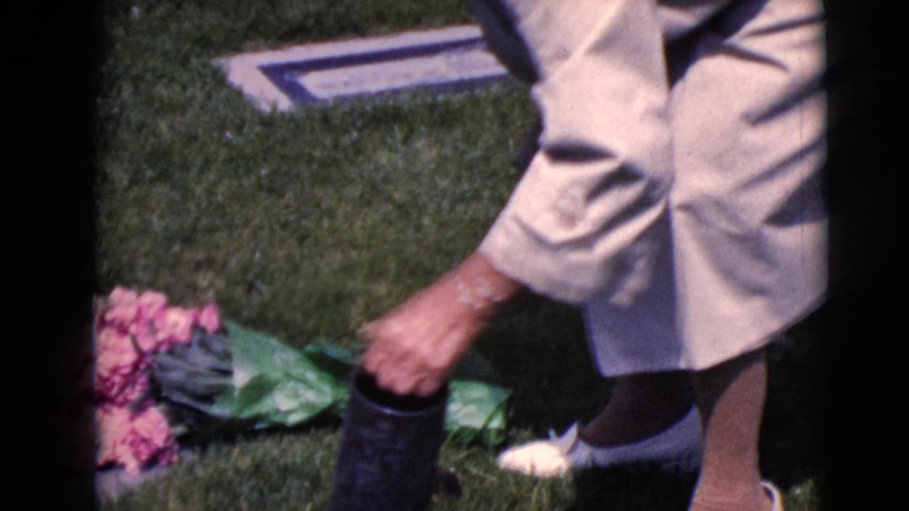 Close view of a woman reaching down near flowers and a dark cane in a cemetery.