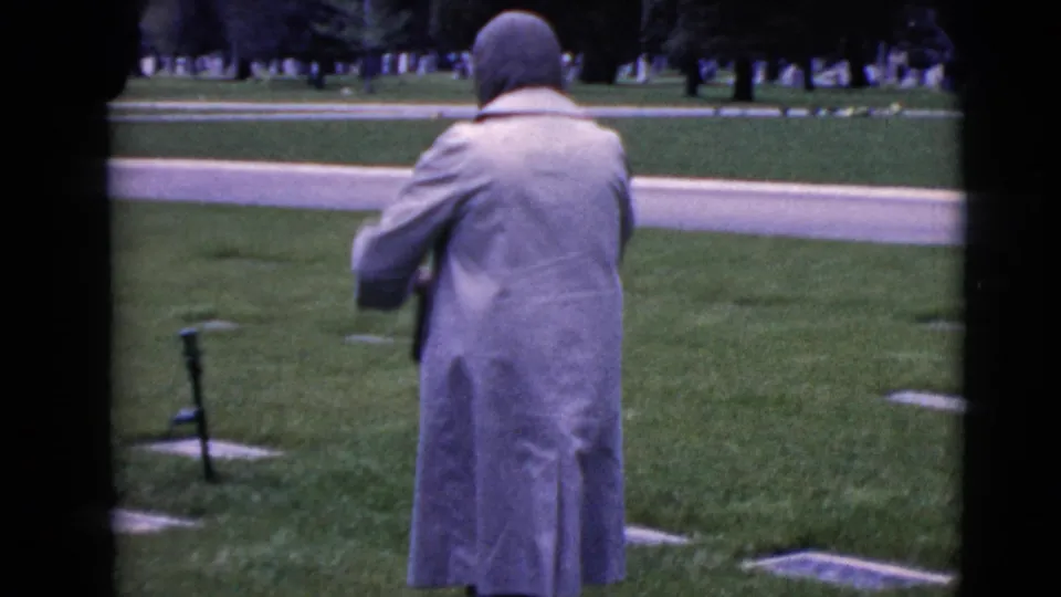 Seen from behind, the woman in a pale coat and head covering stands among flat grave markers in a cemetery setting.