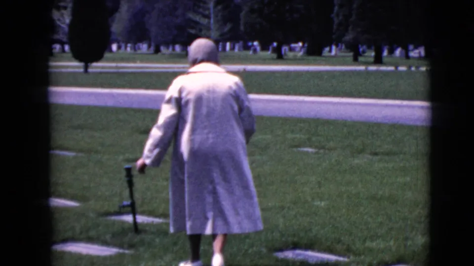 The woman walks away across the cemetery grass, with flat grave markers and a paved road visible behind her.