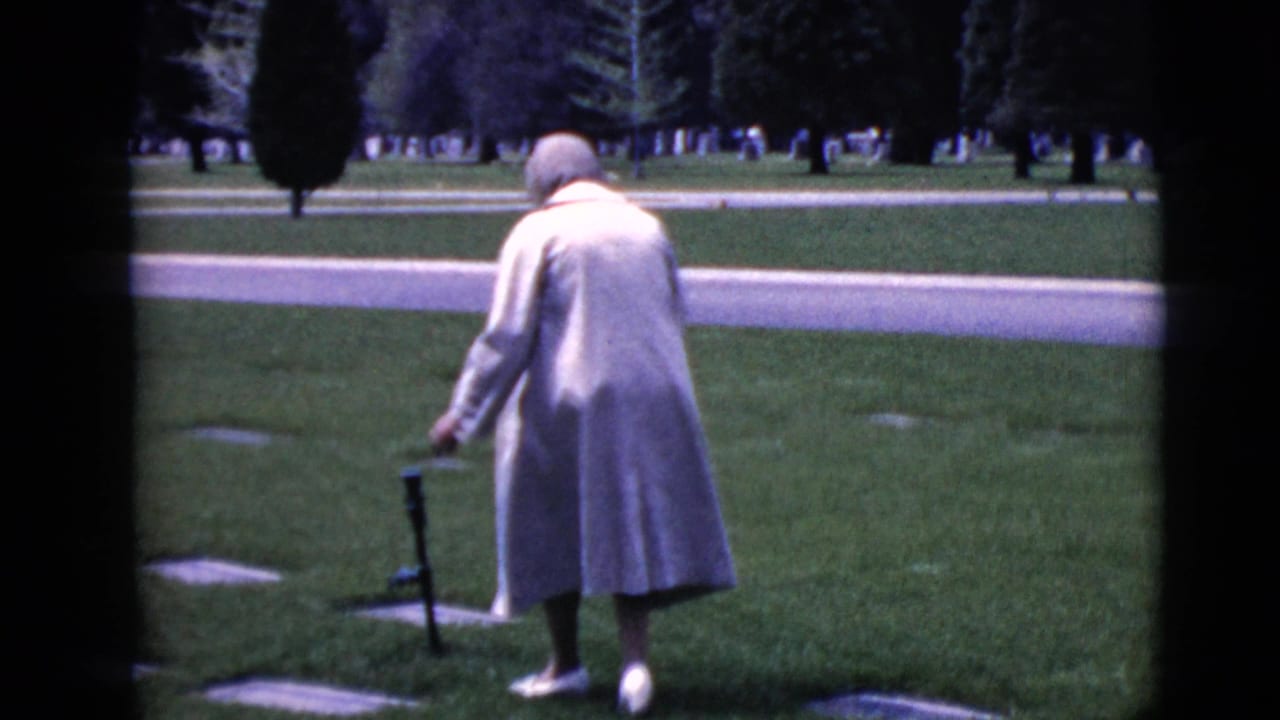 Woman in a light coat walks with a cane across a grassy cemetery.