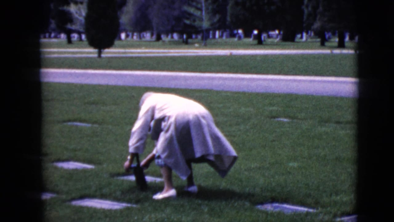 The woman bends carefully near a flat grave marker in the grassy cemetery, with her cane beside her and trees in the background.