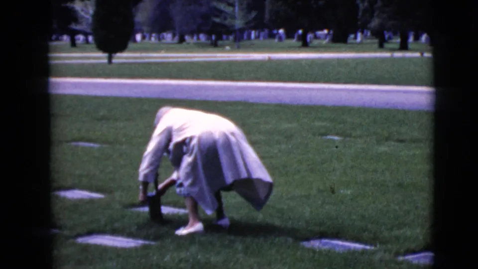 A woman in a light coat remains bent near a flat grave marker, appearing to place or adjust something at the gravesite.