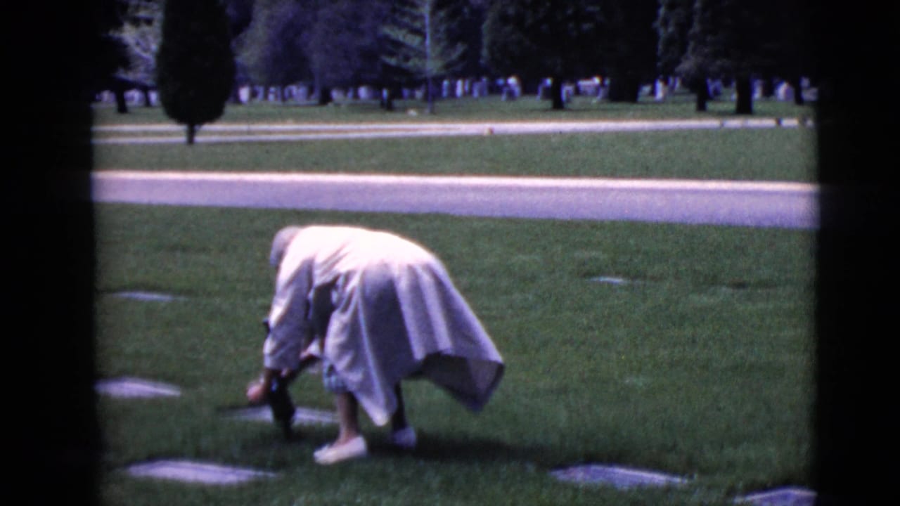 Elderly woman bends over a flat grave marker on a cemetery lawn.