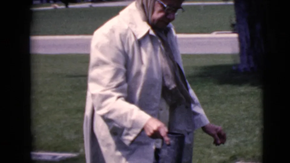 An elderly woman wearing glasses and a pale coat walks through the Clarksdale, Arizona cemetery in 1968 home-movie footage.