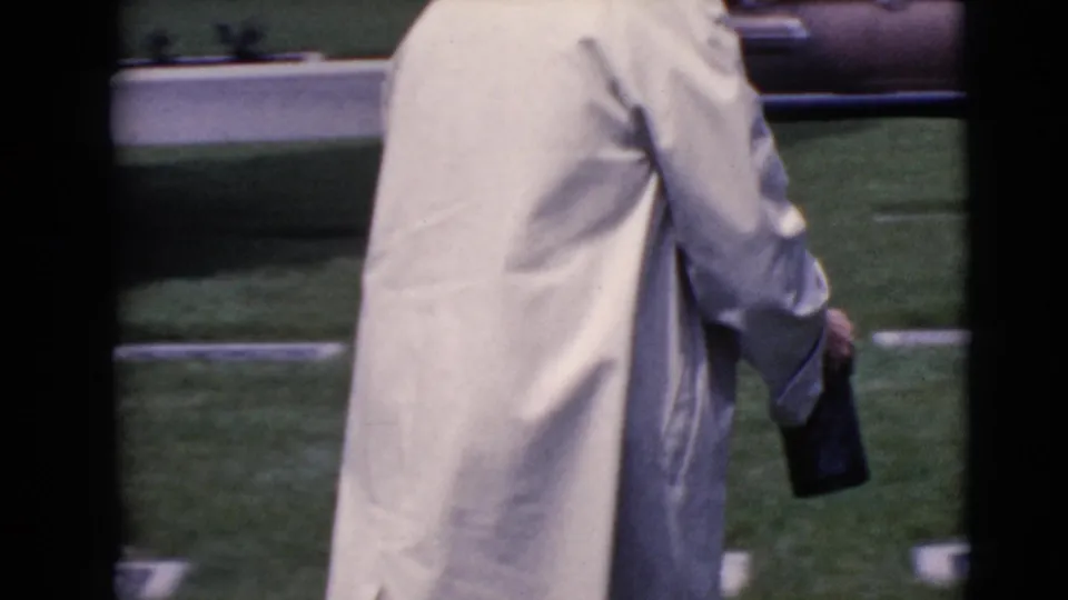 The elderly woman is seen from behind in a light coat, carrying a dark handbag as she walks among cemetery markers in Arizona.