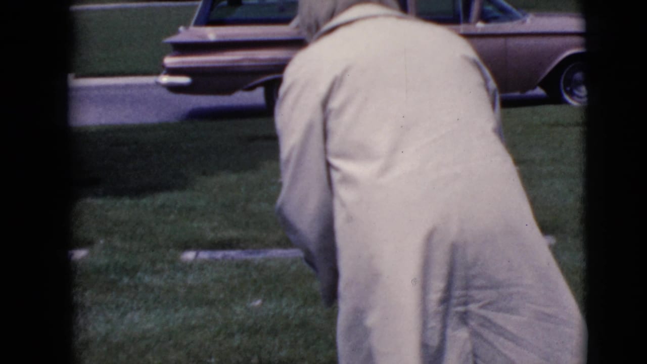 Woman in a pale coat bends over a grassy cemetery near a parked station wagon.