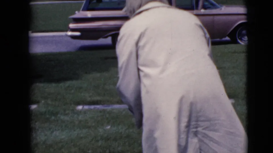 In 1968 Clarksdale, Arizona footage, a woman in a pale coat bends near grave markers with a vintage station wagon in the background.