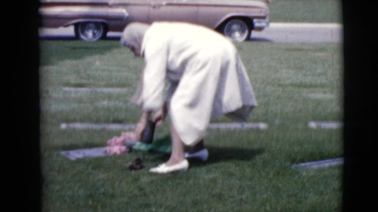 A woman in a light coat and head covering places pink flowers at a flat grave marker in a Clarksdale, Arizona cemetery.