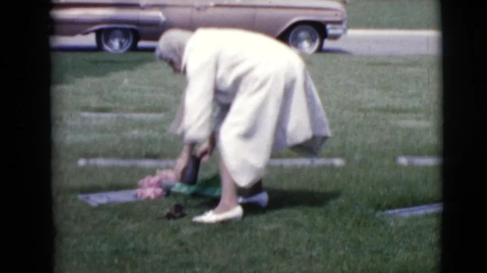 A woman in a light coat and head covering places pink flowers at a flat grave marker in a Clarksdale, Arizona cemetery.