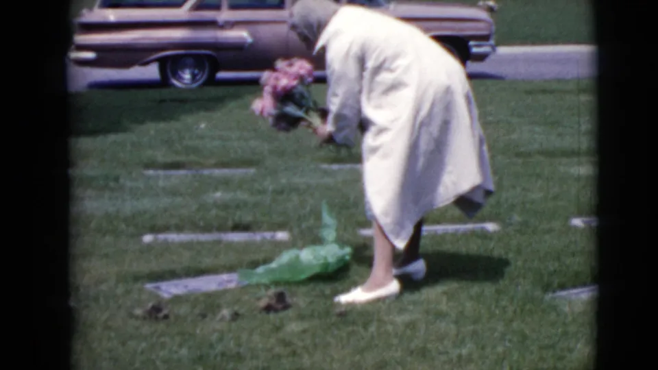 The 1968 home-movie frame shows the woman holding pink flowers near a flat grave marker, with a green bag on the cemetery grass.