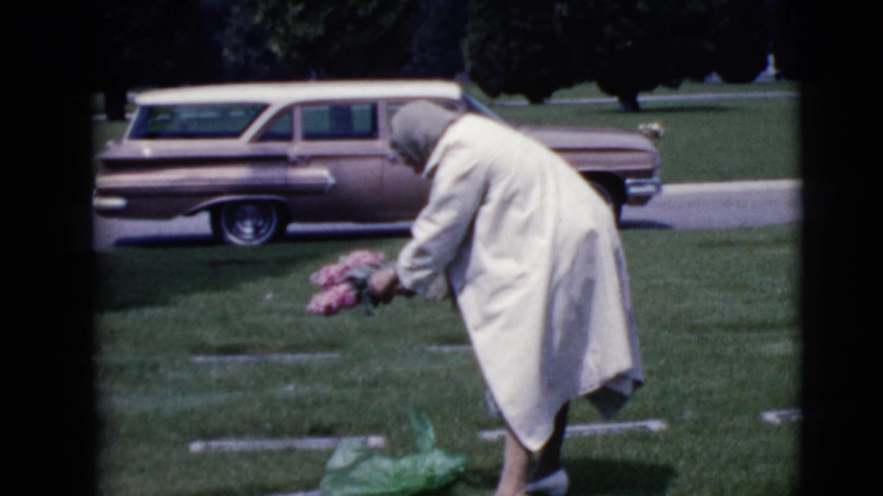 In 1968 Clarksdale, Arizona footage, an older woman in a light coat bends over a grave with pink flowers as a station wagon sits nearby.