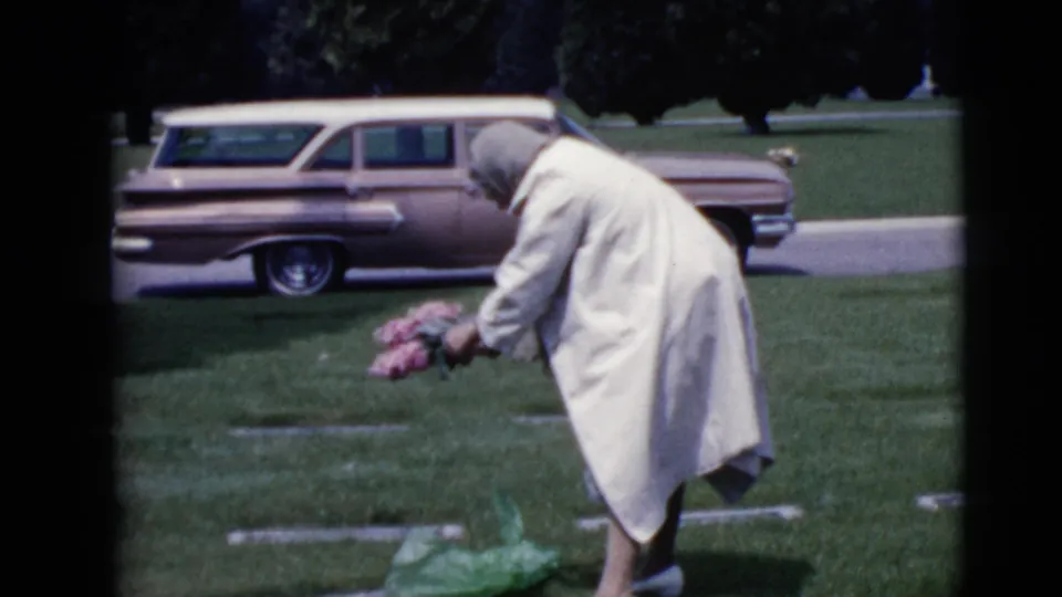 In 1968 Clarksdale, Arizona footage, an older woman in a light coat bends over a grave with pink flowers as a station wagon sits nearby.