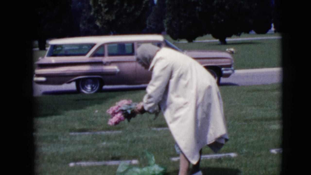 An older woman in a light coat and head covering leans toward a grave with pink flowers in 1968 Clarksdale, Arizona home-movie footage.