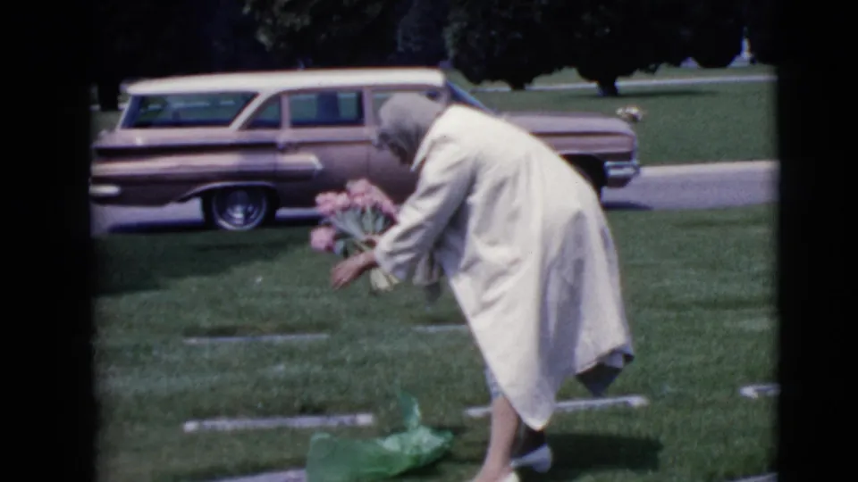 The 1968 home-movie frame shows an older woman adjusting pink flowers near a flat cemetery marker in Clarksdale, Arizona.