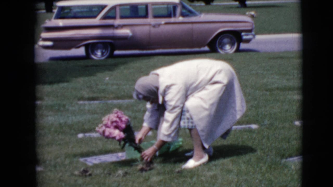 Older woman bends over a flat headstone with pink flowers in a grassy cemetery.