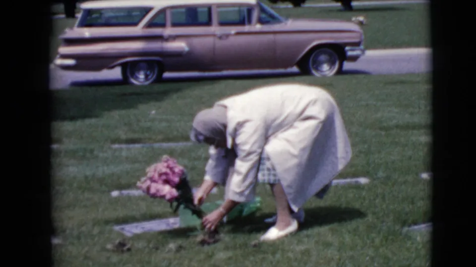 In 1968 Clarksdale, Arizona footage, an older woman in a light coat bends to place pink flowers at a cemetery headstone.