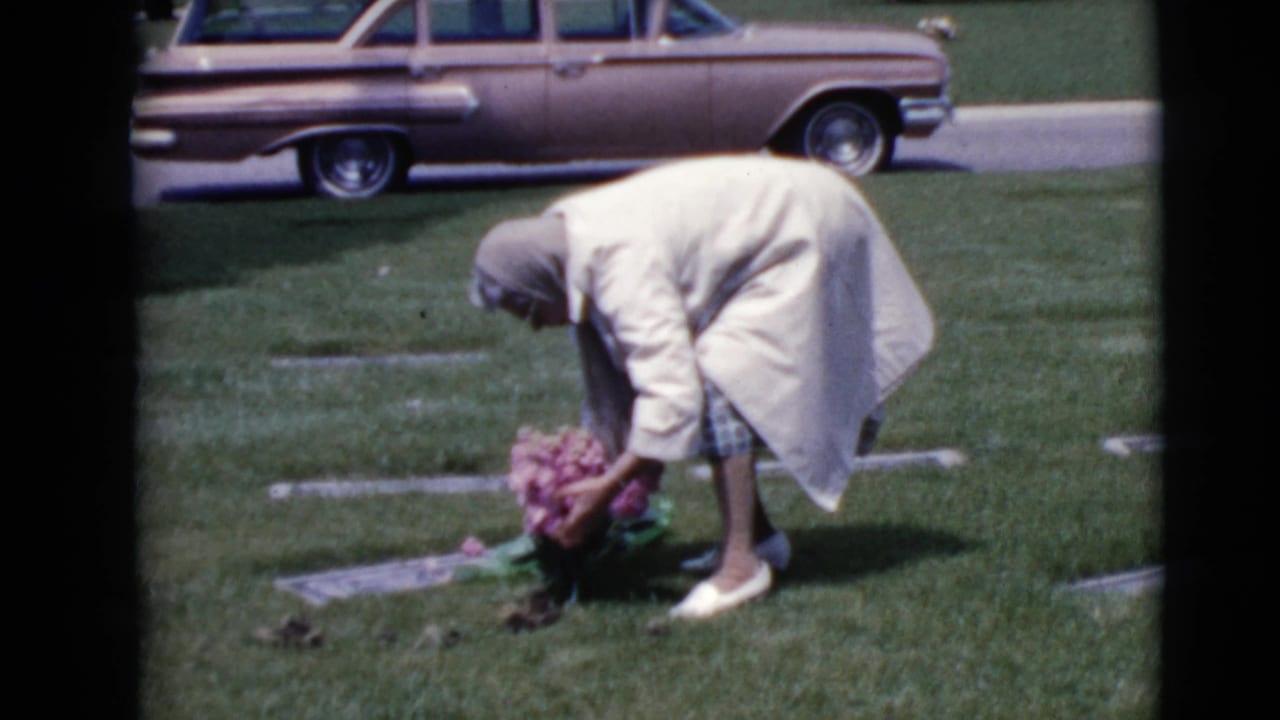 The woman continues arranging pink flowers at a flat grave marker as a 1960s station wagon sits near the cemetery path.