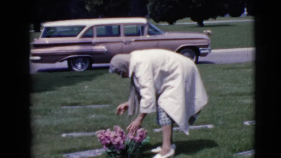 In the cemetery scene, the older woman rises slightly while tending two pink flower clusters near a flat headstone.