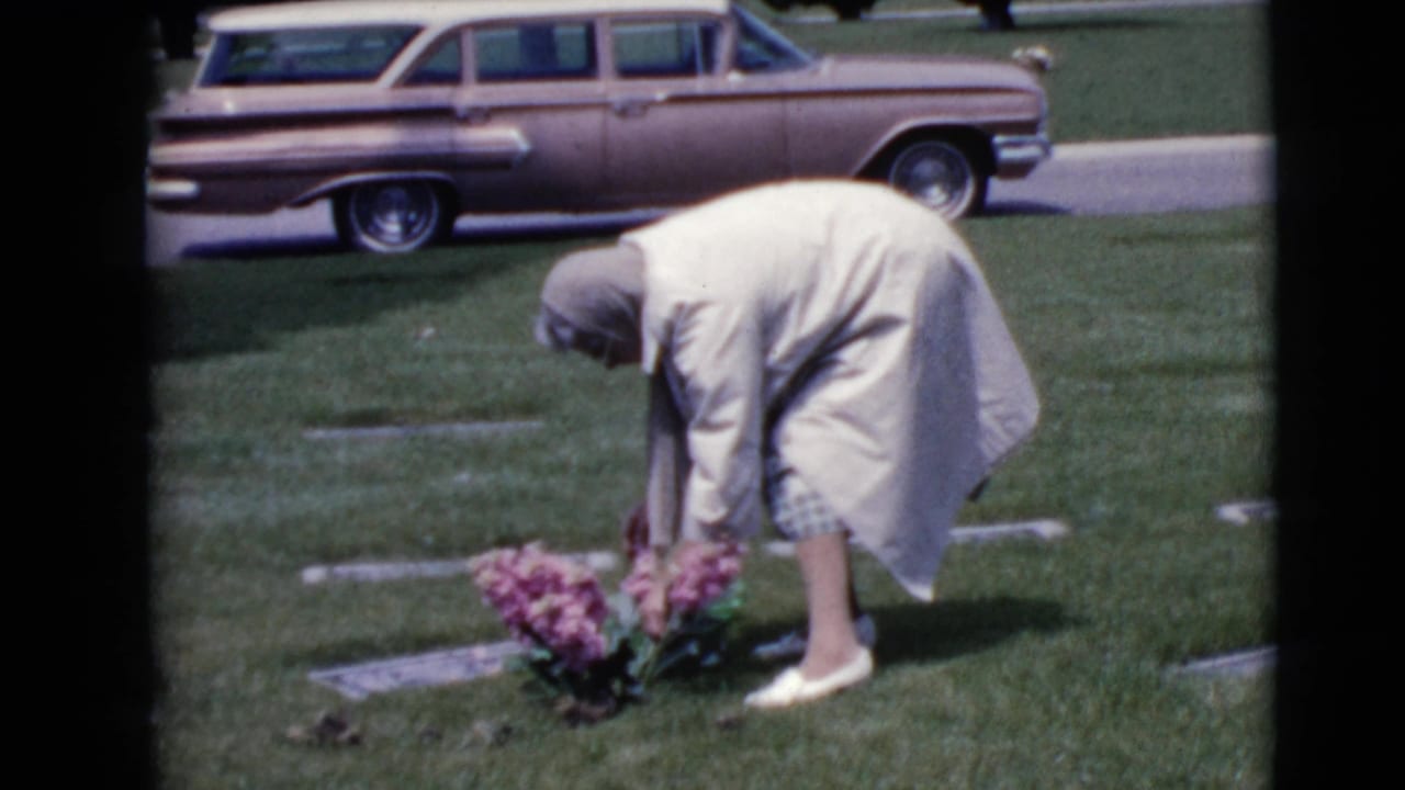 In a 1968 Clarksdale, Arizona cemetery scene, a woman in a light coat bends to arrange pink flowers near flat grave markers.