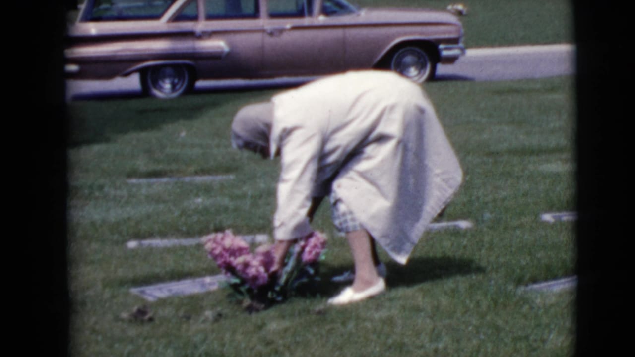 A woman tends pink flowers at a cemetery grave marker while a 1960s station wagon is parked behind her on a paved lane.